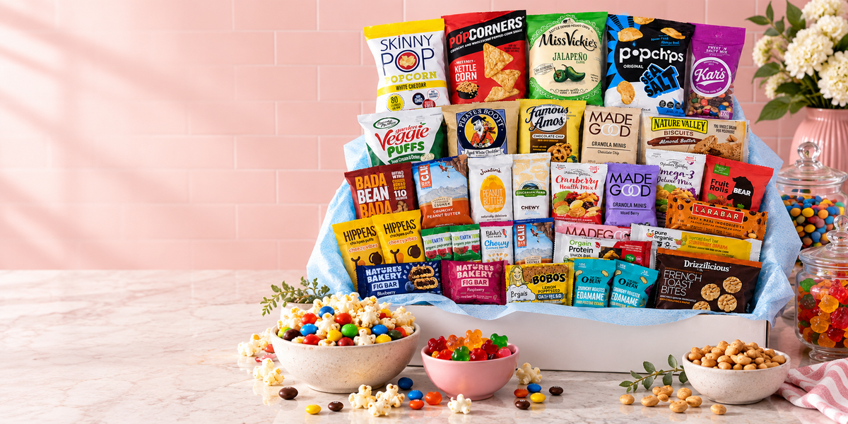 A white gift box lined with pale blue tissue, filled with individually wrapped snack bags and bars, with bowls of popcorn, gummies and peanuts on the counter beside it